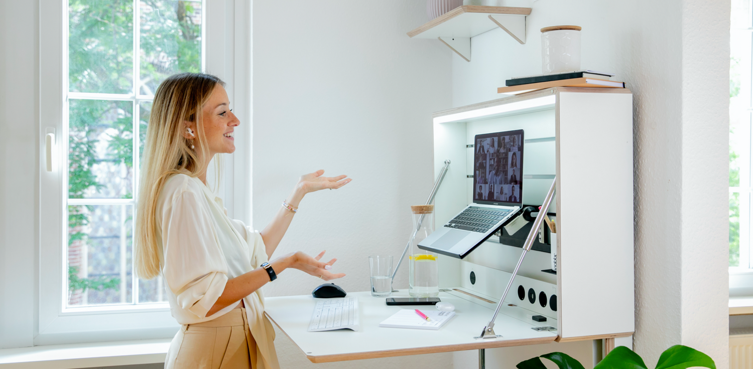 Ein Frau steht an einem weißen Klappt. Schreibtisch und spricht gerade während eines VideoCalls in die Kamera. Sie arbeitet mit der klappbaren Laptopablage und die Tageslichtlampe ist angeschaltet.