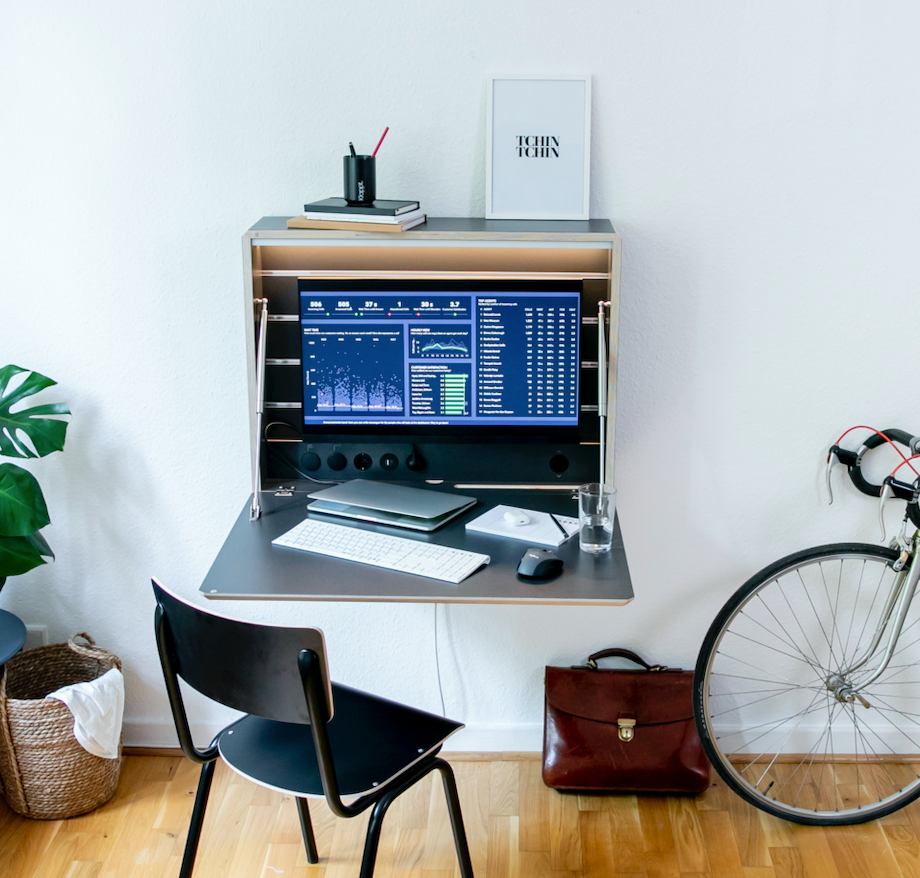 Zu sehen ist der Klappt. Schreibtisch in anthrazit mit einem eingehängten Monitor. Die Arbeitsbeleuchtung ist eingeschaltet und auf der Arbeitsfläche stehen Laptop, Tastatur, Maus, Notizzettel und ein Glas Wasser.