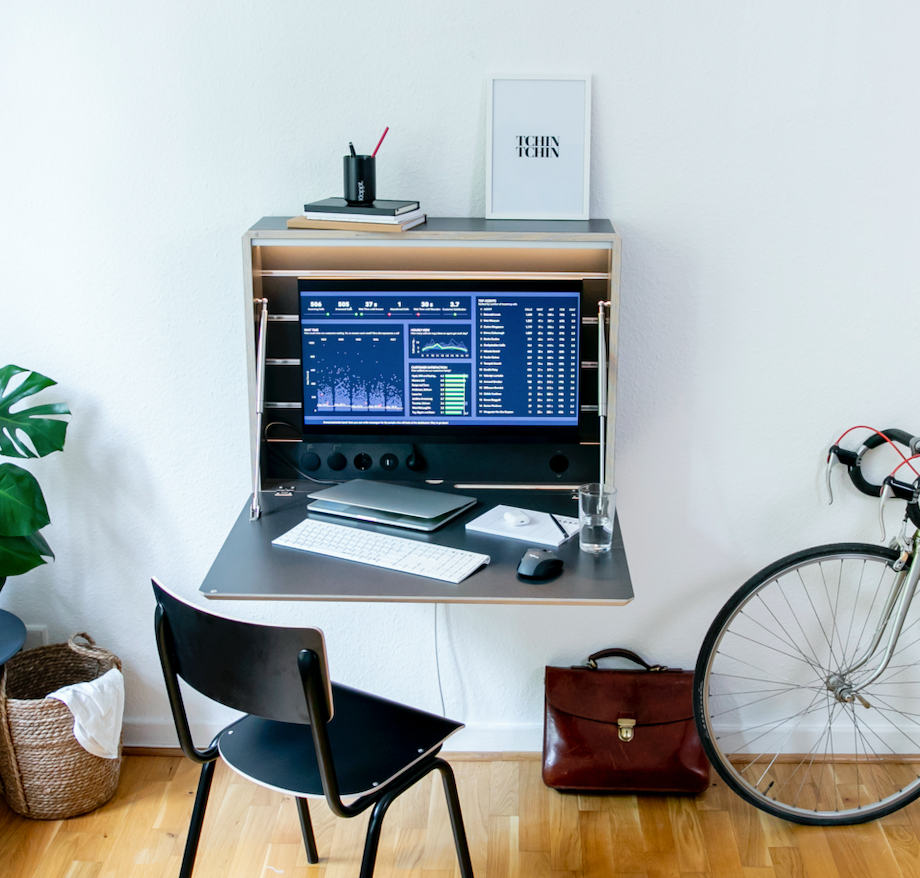 Zu sehen ist der Klappt. Schreibtisch in anthrazit mit einem eingehängten Monitor. Die Arbeitsbeleuchtung ist eingeschaltet und auf der Arbeitsfläche stehen Laptop, Tastatur, Maus, Notizzettel und ein Glas Wasser. 